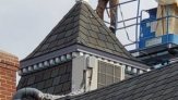 Carpenter stands on roof with hands on the spire as he puts it in place.  The lift is next to him, and his harness is attached via lanyard to the lift.