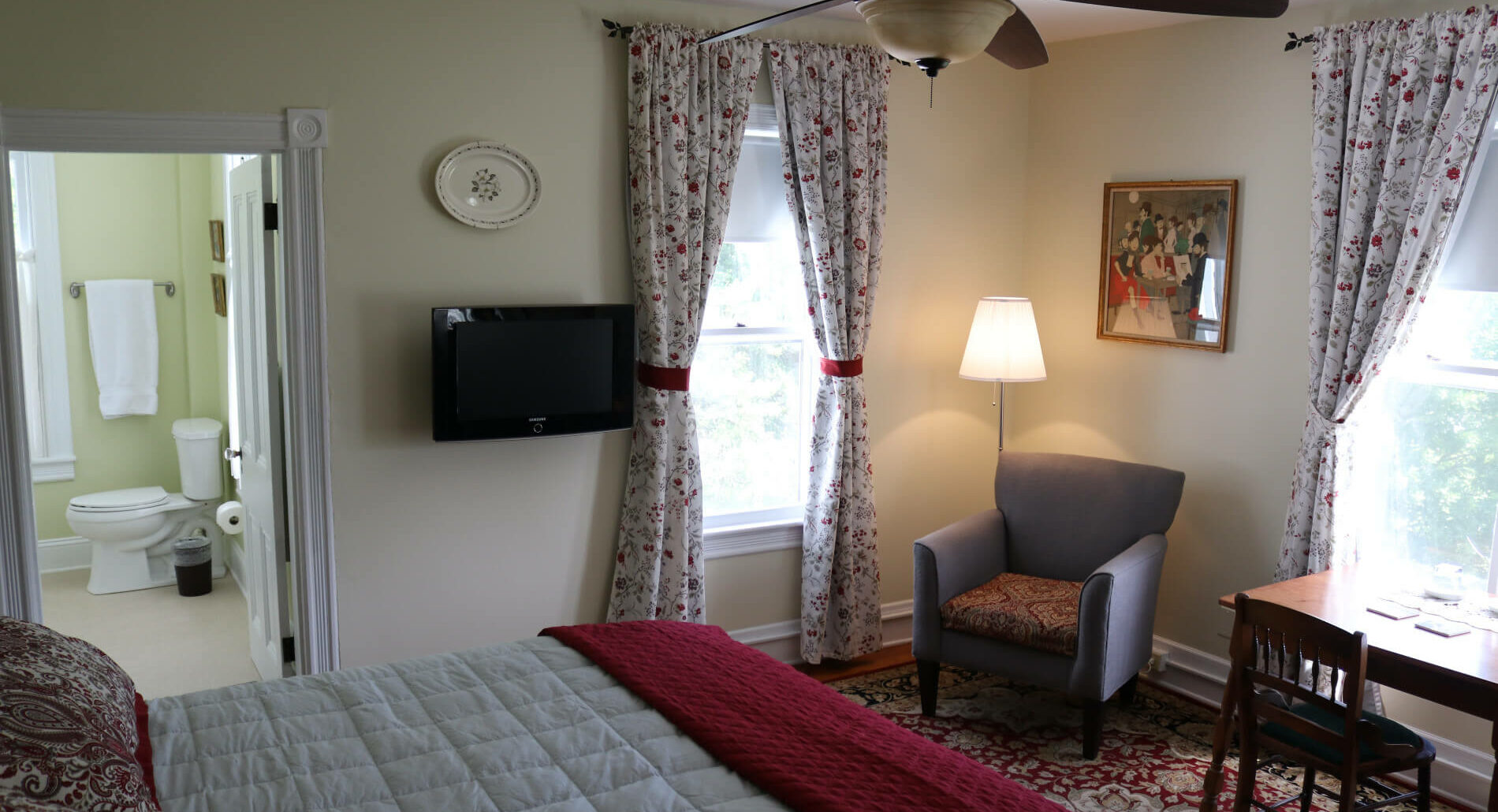Bedroom with two windows with flowered curtains, table, chair, and reading lamp, red rug. Bed in foreground, looking into bathroom.