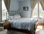 Light blue room with hardwood flooring, two large windows, light blue bedding, bedside table and chair