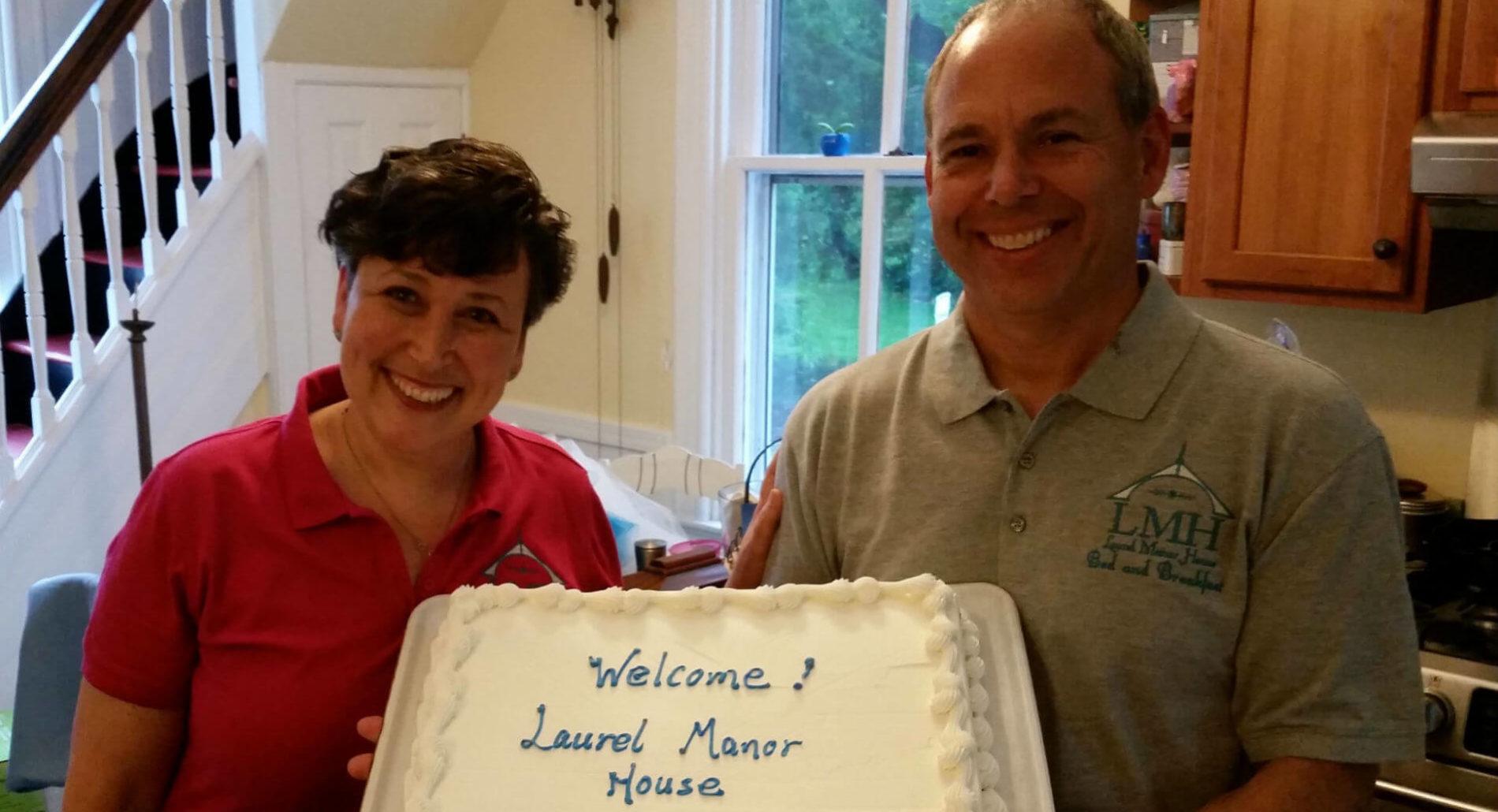 Women in red shirt and man in grey shirt, both smiling, hold cake that says "Welcome to Laurel Manor House".