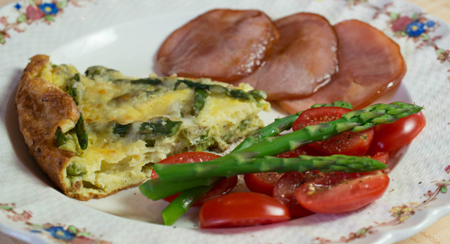 A white plate displaying a quiche, Canadian bacon, bright green asparagus and vibrant red cherry tomato slices