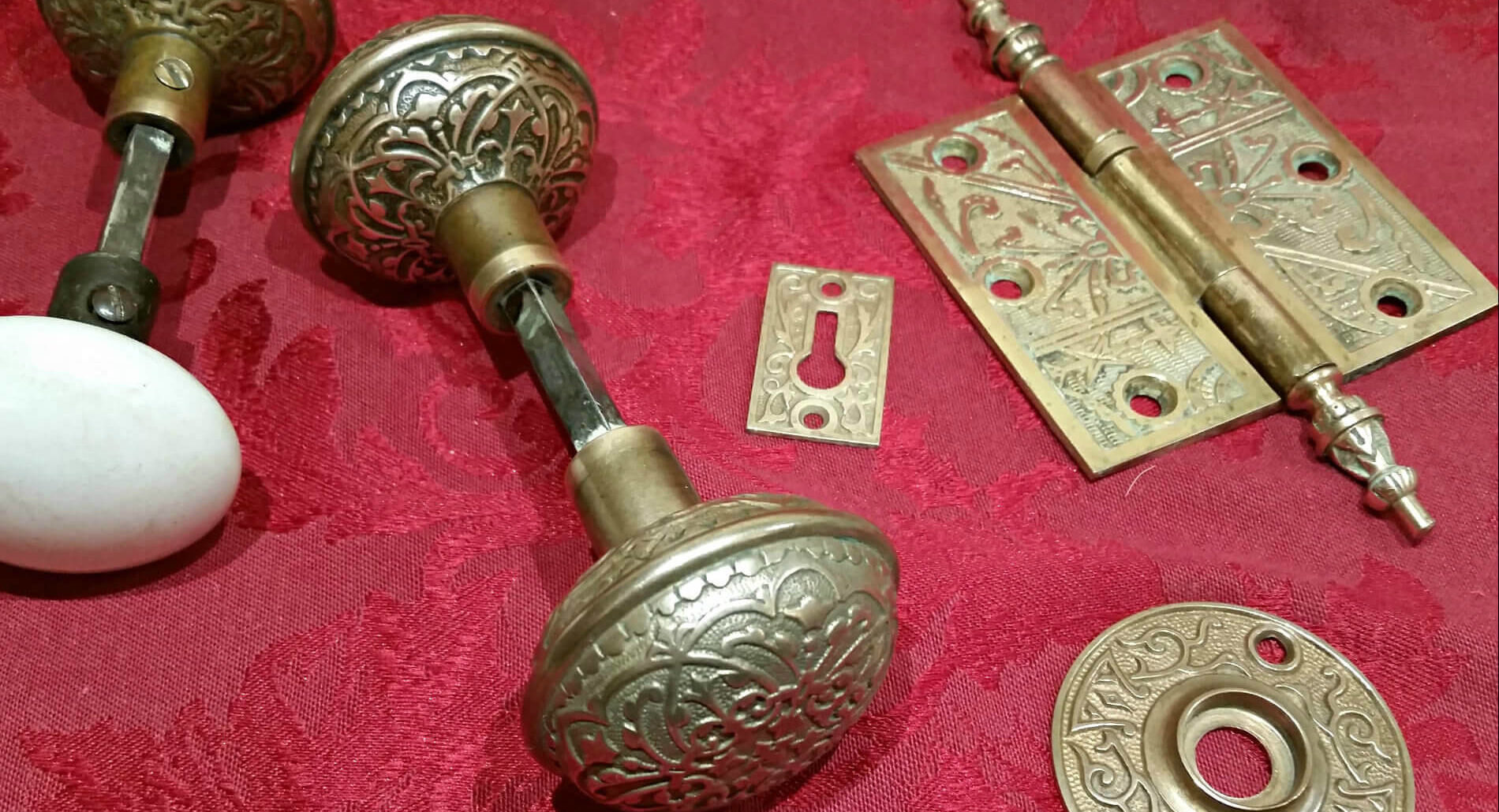 Gold vintage pieces to a door displayed including two doorknobs and a door hinge against a red table cloth