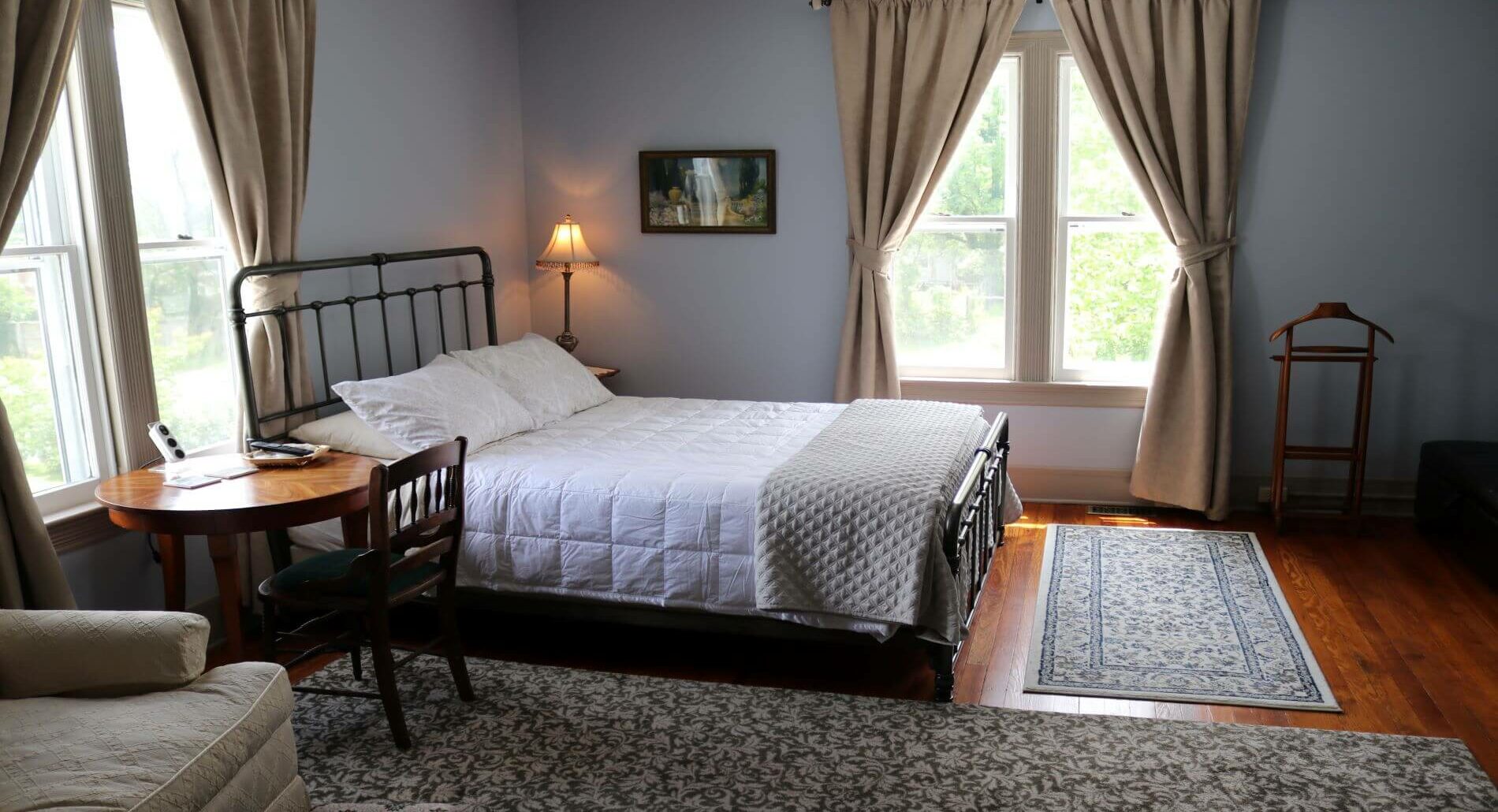 Light blue room with white bedding, two large windows, table and chair and beside table with a lamp