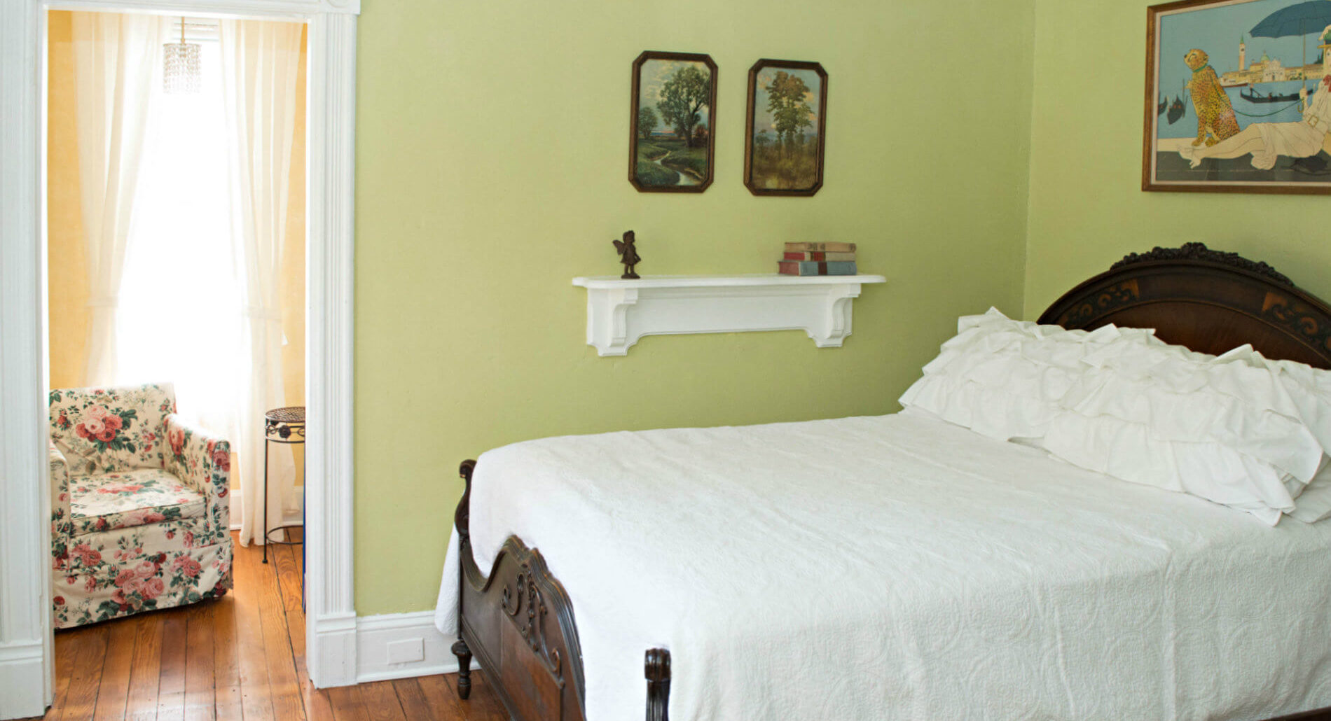 Light green room with white bedding, hardwood flooring, doorway to a yellow sitting room with a floral cushioned chair