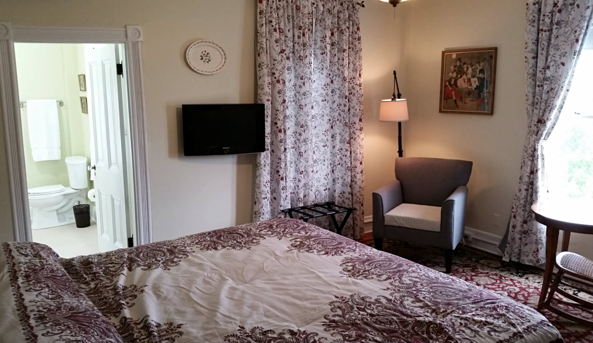 Beige guest room with burgundy and ivory bedding, flat screen tv, a chair and reading lamp