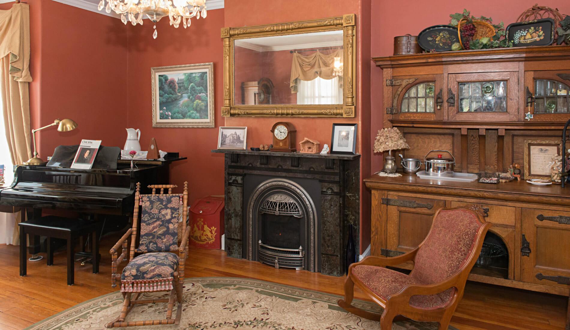 Dark orange room featuring a shiny black grand piano, a fireplace and mantel, and two cushioned sitting chairs