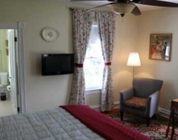 Beige guest room with burgundy and ivory bedding, flat screen tv, a chair and reading lamp