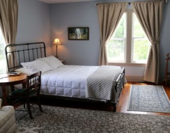 Light blue room with white bedding, two large windows, a vintage writing desk and beside table with a lamp