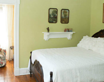 Light green room with white bedding, hardwood flooring, doorway to a yellow sitting room with a floral cushioned chair