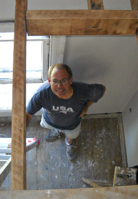 White room undergoing construction, man standing below tan wooden rafters holding onto a ladder