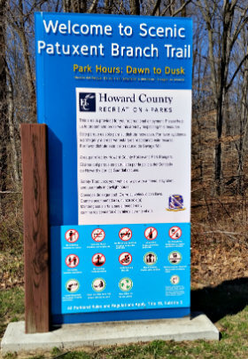 Large exterior blue sign attached to a wooden brown post which says welcome to scenic patuxent branch trail