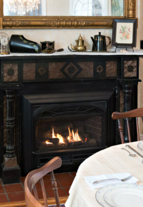 Round brown wooden table with a cream tablecloth surrounded by wooden chairs and a black fireplace and white mantel