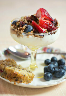 Small glass dish containing a yogurt parfait with granola, blueberries, and bright red strawberries