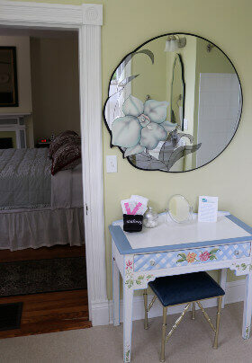 Vanity table with blue seat. Circular mirror with floral design on wall. Through doorway is bedroom; bed has white coverings, red pillows.